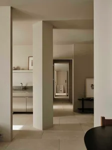 A modern kitchen and hallway area with white surfaces, a sink, refrigerator, and dining table, with natural light and a warm atmosphere