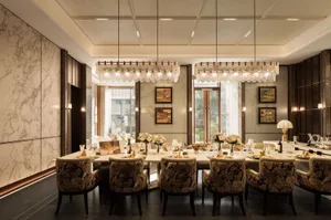A dining room with a long wooden table and chairs, surrounded by wine glasses, cups, and vases, with a large window allowing natural light to enter, creating a warm and inviting atmosphere