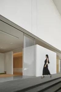 A woman in a black dress walks through a well-lit, modern building with a glass exterior and natural light streaming in from the windows