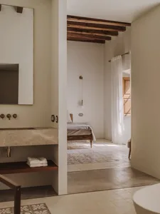 A minimalist bedroom with a bed, a wooden chair, and a white rug, well-lit by natural light from the window