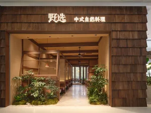 A modern restaurant with a wooden exterior, well-lit interior, and natural materials, featuring furniture, a potted plant, and a welcoming atmosphere