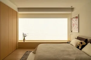 A well-lit bedroom with a bed, window, and vase with flowers, featuring predominantly white materials
