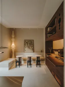 A well-lit living room with a dining area, featuring a dining table, chairs, a couch, bookshelf, vase, and potted plant, in a predominantly white space with a modern appearance