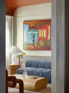 Comfortable living space with a blue couch, wooden furniture, and a glass table, featuring a painting and a potted plant, with natural light from a window