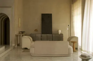A cozy living room with a beige color scheme, featuring a large couch, a chair, and various decorative pieces, with natural light streaming in through the windows