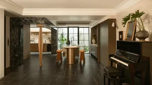 Modern kitchen and dining space with a piano, dining table, and kitchen counter, decorated with plants, vases, and bottles, illuminated by natural light