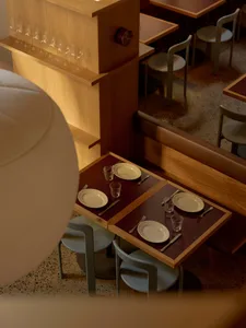 A dimly lit restaurant with wooden floor and tables set with plates, silverware, and glasses, featuring a modern style with wood and metal elements