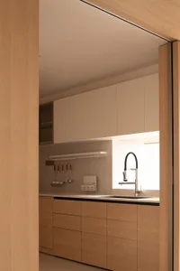 A modern kitchen with a white color scheme, wooden cabinets, a sink on the countertop, and a refrigerator in the background, lit by natural light