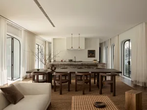 A warm and inviting kitchen and dining area with a large dining table, kitchen counter, and couch, decorated with a vase, bowls, and a potted plant, with natural light streaming in through the windows