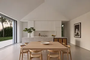 A modern kitchen with a wooden dining table, white cabinets, and natural light from a window, set with a bowl and oranges