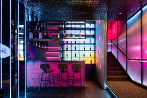A lively bar with a pink and purple color scheme, featuring a counter with stools and numerous bottles on display