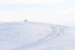 A snowy landscape with a road going up a hill, featuring two people walking and a car in the distance