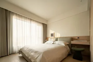 A calm bedroom with a bed, chair, and natural light from a window, featuring a white color scheme