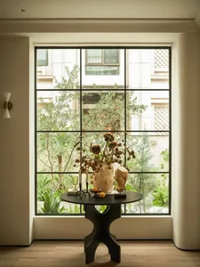 A living room with a table in front of a large window, featuring a vase of flowers and a candle, surrounded by natural light