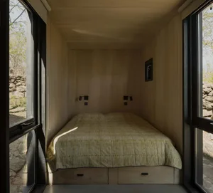 A small bedroom with a bed near a window, allowing natural light to enter, and a door for access to the rest of the space