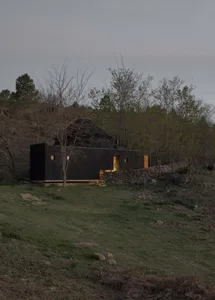 A small, dark wooden cabin with a stone wall, surrounded by trees, featuring a mix of a living room and bedroom with a bed and couch inside
