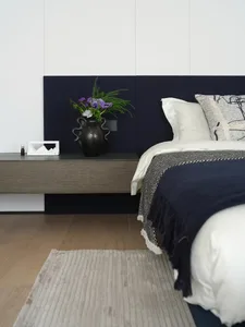 A well-lit bedroom with a bed, nightstand, and vase with flowers, featuring predominantly white materials and a clean appearance