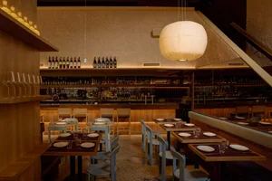 Cozy restaurant with wooden floor, tables, chairs, and a bar, featuring dim lighting and various bottles, creating a warm and intimate atmosphere