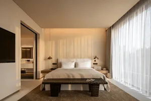 A well-lit hotel bedroom with a bed, bench, and lamp, featuring a white color scheme and natural light from the window