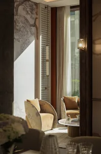 A modern living room with two chairs, a dining table, and a vase, lit by natural light from a large window
