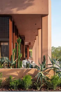 A red brick building with a patio area featuring cacti in pots, chairs, and a dining table, surrounded by natural sunlight and a warm atmosphere