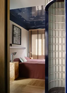 A modern bedroom with a blue color scheme, a bed, nightstand, and picture on the wall, with natural light streaming in through the open door