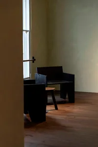 Black and white photograph of a minimalist living space with a wooden floor, a black chair, and a black bench, featuring a window in the background
