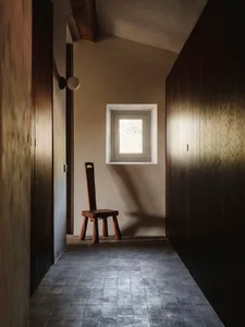 A small, dark room with a wooden floor and a chair in the center, lit by a window on the wall