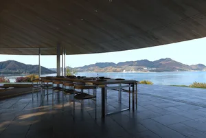 A well-lit dining room with a wooden table and chairs, surrounded by a large window with a view of the outdoors and a body of water