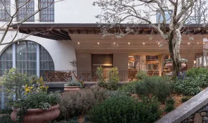 A rustic restaurant with a wooden exterior, featuring a large wooden structure and a covered outdoor area with dining tables and chairs, bathed in natural sunlight