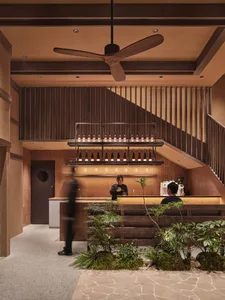 Warm and inviting restaurant interior with a bar, staircase, and potted plants, featuring wood accents and a large fan