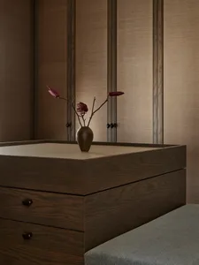 A cozy bedroom with a wooden dresser, a vase of flowers, and a bed in the background, creating a warm and inviting atmosphere