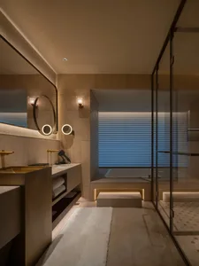 A modern bathroom with a beige color scheme, featuring a large mirror, sink, and bathtub, with a dimly lit atmosphere and a combination of natural and artificial light sources