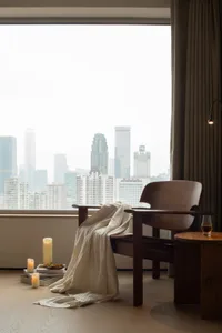 Cozy living room with a wooden chair near a window, a table in front, and a city view through the large window