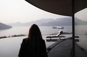 一個女人站在玻璃陽台上,眺望著大片的水域,遠方可見一艘船,置身在現代豪宅中