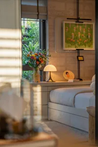 A minimalist bedroom with a white bed, natural light from a window, and additional lighting from a lamp, creating a clean and inviting space