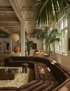 A warm and inviting living room with a brown sectional sofa, dining table, and various decorative items, surrounded by potted plants and well-lit
