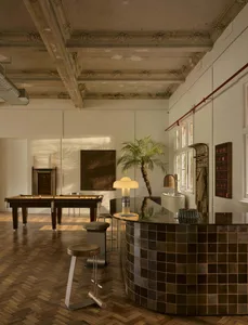 A modern living room with a pool table, bar, and dining table, featuring a tile floor, wood furniture, and a potted plant, with a well-lit and inviting atmosphere