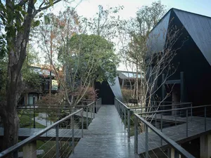 A modern black house with a wooden walkway and metal railing, surrounded by trees, with natural sunlight and a bright atmosphere
