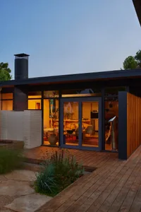 A modern house with a large open living room and kitchen, featuring a fireplace, couch, dining table, and refrigerator, with a black exterior and wooden deck, well-lit for a warm atmosphere