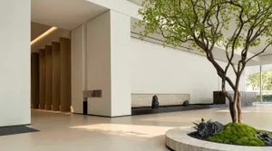 A modern building with a white exterior, featuring a large fireplace, a tree in the courtyard, and a stone wall, blending indoor and outdoor spaces with bright lighting