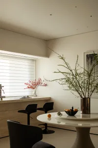 A kitchen with a dining table, chairs, and a potted plant, featuring a sink, refrigerator, and natural light from the windows