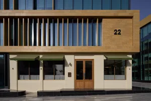 A tan-colored building with a wooden door and a number 22 on it, set against a natural outdoor backdrop with a bench in the foreground