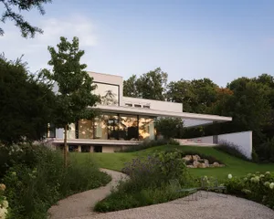 A serene residential house with a white exterior, large window, and patio area, surrounded by a garden with a tree and bushes
