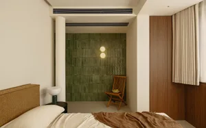 A modern bedroom with a green tiled wall, white floor, and a bed, chair, and light, illuminated by natural sunlight through a window