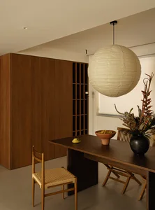 A modern dining area with a wooden table, chairs, and a hanging light, featuring a bowl and a plant in a vase