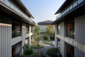 A serene outdoor living space with a mix of gray and brown tones, featuring a large courtyard with plants, trees, and a central rock, bathed in natural daylight