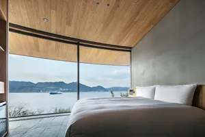 A serene bedroom with a large bed, white bedding, and a view of the ocean through a window, bathed in natural light