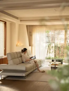 A woman sits on a white couch in a well-lit living room with a chair and dining table, creating a comfortable atmosphere