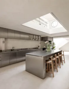 A well-lit kitchen with a large steel island, wooden chairs, and a potted plant, surrounded by white walls and a steel sink